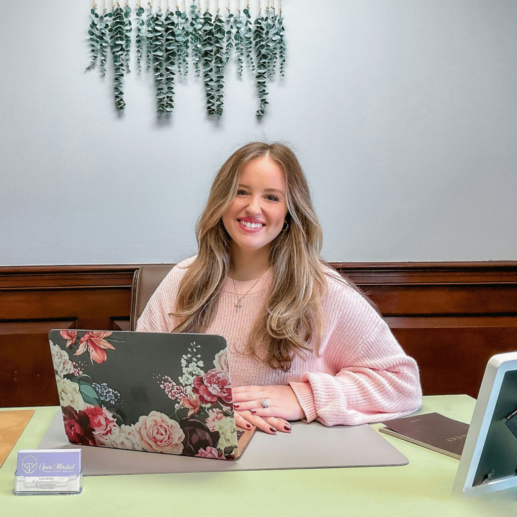 Image of Alexa Sadowsky, owner of Open Minded Marriage and Family Therapy, PLLC, smiling as she sits at her desk with her laptop open and her hands folded softly in front of her.