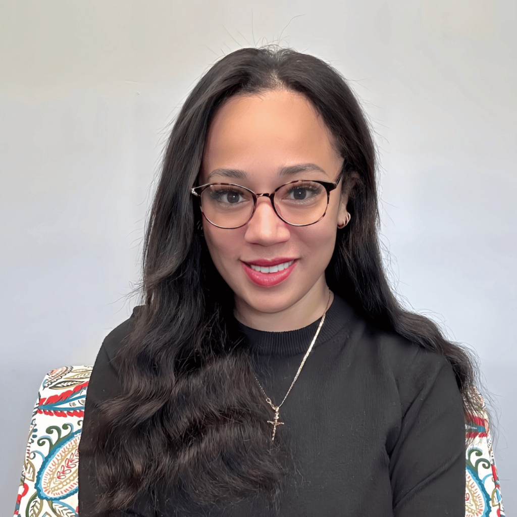 Image of Novelle A. Court with long black hair and a black top, sitting in a chair as she smiles at the camera. She wears dark rimmed glasses.